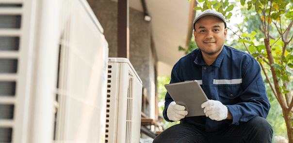 A technician wearing a cap, gloves, and uniform kneels outdoors beside air conditioning units, holding a tablet and smiling at the camera.