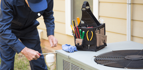 A technician in uniform uses a screwdriver to repair an outdoor air conditioning unit, with a tool bag and gloves placed on top of the unit.