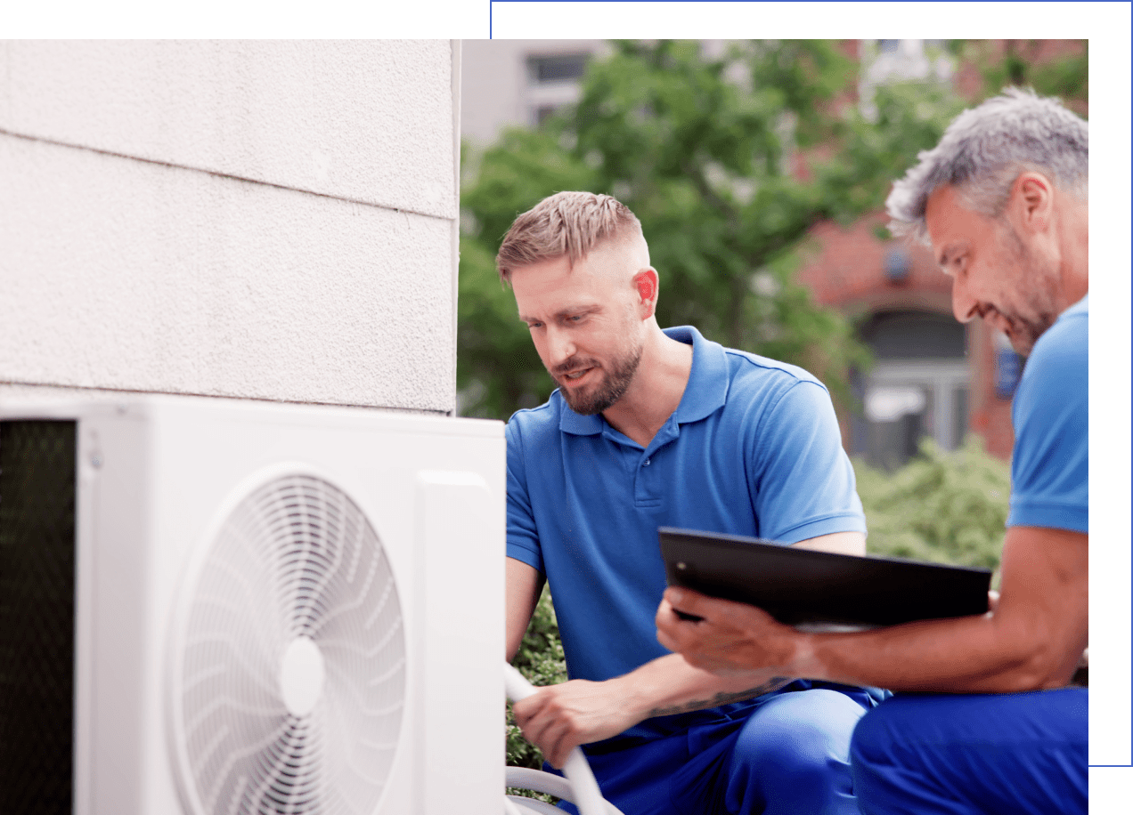 Two men in blue uniforms inspect and discuss an outdoor air conditioning unit; one holds a clipboard, the other examines the unit.